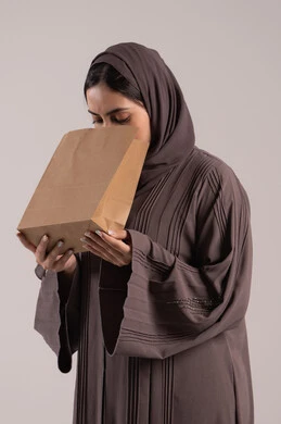 إيماءات التقيؤ والغثيان، الإحساس بالمغص والوجع، الحاجه للمساعدة وطلب العلاج، بورتريه مقرب من الجانب لامرأة عربية خليجية إماراتية ترتدي العباءة تحمل بيدها كيس ورقي، شابة سعودية مريضة، خلفية بنية