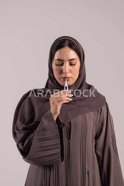 شعور بالتعب المصحوب بالحمى، وضع خطة علاجية، الحاجة للمساعدة وزيارة الطبيب، الإحساس بالمرض والإرهاق، بورتريه لامرأة عربية خليجية اماراتية محجبة ترتدي عباءة تقوم بقياس حرارتها باستخدام الميزان الرقمي، شابة سعودية مريضة، خلفية بنية