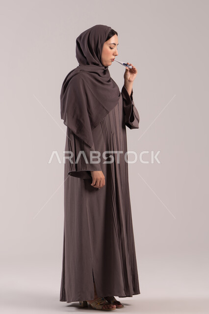 ارتفاع درجة الحرارة والتعب الجسدي، وضع خطة علاجية، الحاجة للمساعدة وزيارة الطبيب، الإحساس بالمرض والإرهاق، بورتريه من الجانب لامرأة عربية خليجية اماراتية محجبة ترتدي عباءة تقوم بقياس حرارتها باستخدام الميزان الرقمي، شابة سعودية مريضة، الجسم بالطول الكامل, خلفية بنية