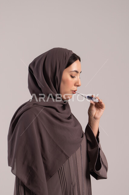 الإصابة بالحمى والتعب، وضع خطة علاجية، الحاجة للمساعدة وزيارة الطبيب، الإحساس بالمرض والإرهاق، بورتريه من الجانب لامرأة عربية خليجية اماراتية محجبة ترتدي عباءة تقوم بقياس حرارتها باستخدام الميزان الرقمي، شابة سعودية مريضة، خلفية بنية