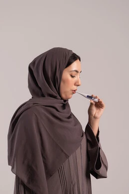 الإصابة بالحمى والتعب، وضع خطة علاجية، الحاجة للمساعدة وزيارة الطبيب، الإحساس بالمرض والإرهاق، بورتريه من الجانب لامرأة عربية خليجية اماراتية محجبة ترتدي عباءة تقوم بقياس حرارتها باستخدام الميزان الرقمي، شابة سعودية مريضة، خلفية بنية