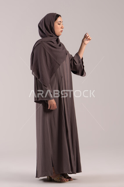 الحاجة للفحص الطبي ووضع برنامج علاجي, الشعور بالتعب والمرض, اتباع إجراءات السلامة والوقاية، الإصابة بالحمى والتعب، بورتريه من الجانب لامرأة عربية خليجية اماراتية محجبة ترتدي عباءة تحمل بيدها ميزان حرارة رقمي، شابة سعودية مريضة، الجسم بالطول الكامل, خلفية بنية