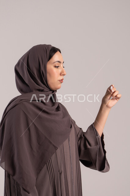 التعب العام المصحوب بالمرض, اتباع إجراءات السلامة والوقاية، الحاجة لزيارة الطبيب ووضع خطة علاجية، الإصابة بالحمى والتعب، بورتريه من الجانب لامرأة عربية خليجية اماراتية محجبة ترتدي عباءة تحمل بيدها ميزان حرارة رقمي، شابة سعودية مريضة، خلفية بنية