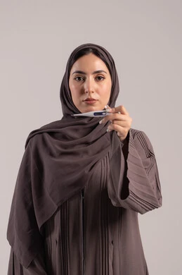 الإرهاق الجسدي والشعور بالمرض, اتباع إجراءات السلامة والوقاية، الحاجة لزيارة الطبيب ووضع خطة علاجية، الإصابة بالحمى والتعب، بورتريه لامرأة عربية خليجية اماراتية محجبة ترتدي عباءة تحمل بيدها ميزان حرارة رقمي، شابة سعودية مريضة، خلفية بنية