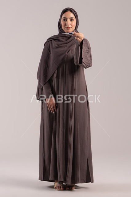 الإحساس بالإرهاق الجسدي والمرض, اتباع إجراءات السلامة والوقاية، الحاجة لزيارة الطبيب ووضع خطة علاجية، الإصابة بالحمى والتعب، بورتريه لامرأة عربية خليجية اماراتية محجبة ترتدي عباءة تحمل بيدها ميزان حرارة رقمي، شابة سعودية مريضة، الجسم بالطول الكامل, خلفية بنية