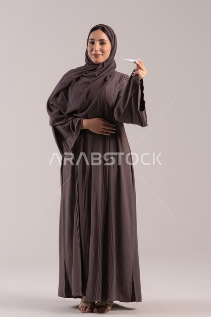 الحاجة للمساعدة وزيارة الطبيب، الإصابة بالحمى والتعب، وضع خطة علاجية، الإحساس بالمرض والإرهاق، بورتريه لامرأة عربية خليجية اماراتية محجبة ترتدي عباءة تحمل بيدها ميزان حرارة رقمي، شابة سعودية مريضة، صورة لكامل الجسم، خلفية بنية