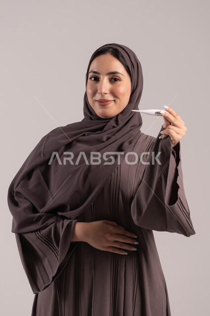 الإصابة بالحمى والتعب, الحاجة للمساعدة وزيارة الطبيب، وضع خطة علاجية، الإحساس بالمرض والإرهاق، بورتريه لامرأة عربية خليجية اماراتية محجبة ترتدي عباءة تحمل بيدها ميزان حرارة رقمي، شابة سعودية مريضة، خلفية بنية