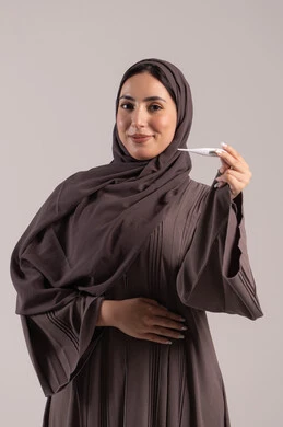الإصابة بالحمى والتعب, الحاجة للمساعدة وزيارة الطبيب، وضع خطة علاجية، الإحساس بالمرض والإرهاق، بورتريه لامرأة عربية خليجية اماراتية محجبة ترتدي عباءة تحمل بيدها ميزان حرارة رقمي، شابة سعودية مريضة، خلفية بنية