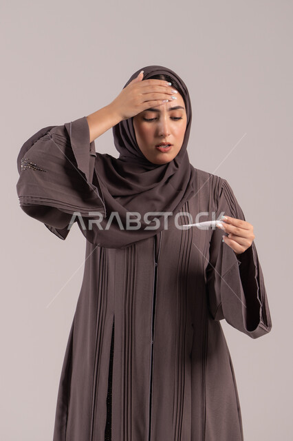الحاجة للمساعدة وزيارة الطبيب، الإصابة بالحمى والتعب، الإحساس بالمرض والإرهاق، بورتريه لامرأة عربية خليجية اماراتية محجبة ترتدي عباءة سوداء تضع يدها على جبينها تقوم بقياس حرارتها باستخدام الميزان الرقمي، وضع خطة علاجية، شابة سعودية مريضة، خلفية بنية