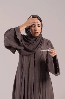 الحاجة للمساعدة وزيارة الطبيب، الإصابة بالحمى والتعب، الإحساس بالمرض والإرهاق، بورتريه لامرأة عربية خليجية اماراتية محجبة ترتدي عباءة سوداء تضع يدها على جبينها تقوم بقياس حرارتها باستخدام الميزان الرقمي، وضع خطة علاجية، شابة سعودية مريضة، خلفية بنية