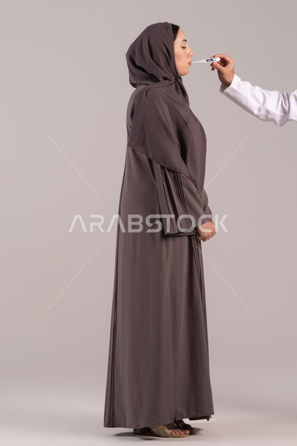 وضع خطة علاجية, الإحساس بالمرض والإرهاق، الإصابة بالحمى والتعب، بورتريه من الجانب لامرأة عربية خليجية اماراتية محجبة ترتدي عباءة  تقوم بقياس حرارتها باستخدام الميزان الرقمي، الحاجة للمساعدة وزيارة الطبيب، شابة سعودية مريضة،  الجسم بالطول الكامل, خلفية بنية