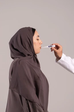 الإحساس بالمرض والإرهاق، الإصابة بالحمى والتعب، بورتريه من الجانب لامرأة عربية خليجية اماراتية محجبة ترتدي عباءة  تقوم بقياس حرارتها باستخدام الميزان الرقمي، الحاجة للمساعدة وزيارة الطبيب، وضع خطة علاجية، شابة سعودية مريضة، خلفية بنية