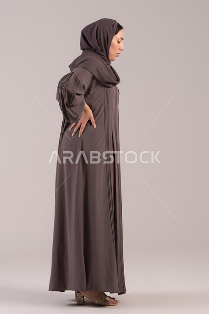 Expressions of pain and backache, feeling ill and fatigued, a side portrait of a veiled Emirati Gulf Arab woman wearing an abaya, placing her hand on her back with gestures of exhaustion, the need for treatment and medical consultation, a sick young Saudi woman, full-body, brown background.