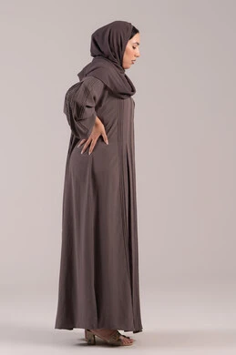 Expressions of pain and backache, feeling ill and fatigued, a side portrait of a veiled Emirati Gulf Arab woman wearing an abaya, placing her hand on her back with gestures of exhaustion, the need for treatment and medical consultation, a sick young Saudi woman, full-body, brown background.