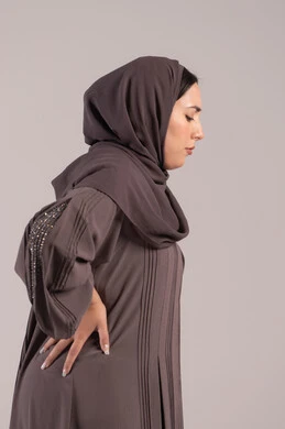 الشعور بالمرض والإرهاق، تعابير الألم ووجع الظهر، بورتريه من الجانب لامرأة عربية خليجية اماراتية محجبة ترتدي عباءة تضع يدها على ظهرها بايماءات التعب، الحاجة للعلاج والاستشارة الطبية، شابة سعودية مريضة، خلفية بنية