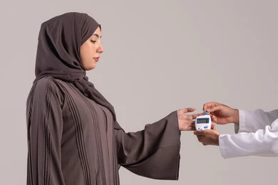 قياس معدل السكر في الدم عبر أجهزة منزلية، بورتريه مقرب ليد ممرض يرتدي اليونيفورم يقوم بفحص نسبة السكر في الدم لمريضة عربية خليجية إماراتية ترتدي العباءة، شركات الرعاية الصحية المنزلية، فحص طبي سريع في البيت، خلفية بنية
