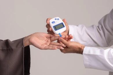 متابعة نسبة السكر في الدم باستخدام أجهزة منزلية، بورتريه مقرب ليد ممرض يرتدي اليونيفورم يقوم بفحص نسبة السكر في الدم لمريضة عربية خليجية إماراتية ترتدي العباءة، شركات الرعاية الصحية المنزلية، فحص طبي سريع في البيت، خلفية بنية