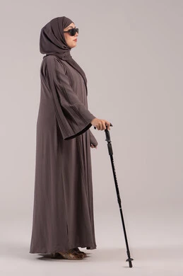The concept of health and illness, loss of vision and sight, the complete reliance of a blind person on a cane in all their movements, a side portrait of a blind Emirati Gulf Arab woman wearing sunglasses and holding a cane, full body, brown background.