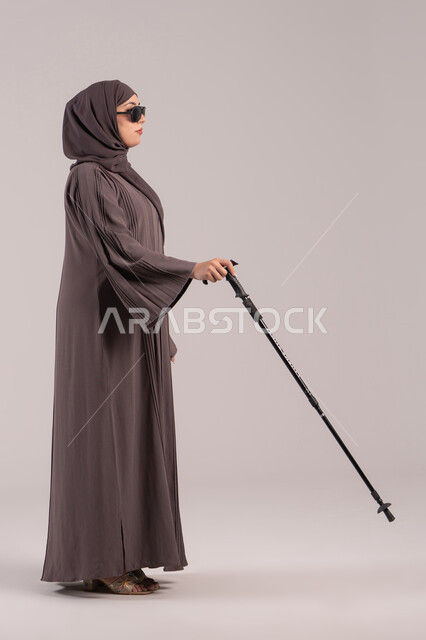 اعتماد الشخص الضرير على العصا بشكل كامل في جميع تحركاته, فقدان الرؤية والبصر, مفهوم الصحة والمرض, بورتريه من الجانب لامرأة عربية خليجية اماراتية كفيفه مصابة بالعمى ترتدي النظارة الشمسية و بيدها العكاز، الجسم بالطول الكامل, خلفية بنية