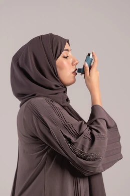 Narrowness and difficulty in breathing, a sick young Saudi woman, maintaining an open airway, a side portrait of a veiled Emirati Gulf Arab woman wearing an abaya holding an asthma inhaler in her hand, inhaling cough medicine, brown background.