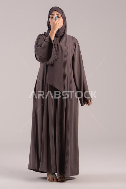 Maintaining airways, a sick young Saudi woman, respiratory problems, a portrait of a veiled Emirati Gulf Arab woman wearing an abaya holding an asthma inhaler in her hand, inhaling cough medicine, full-body image, brown background.