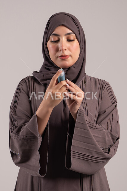 معاناة من مشاكل صدرية وتنفسية, شابة سعودية مريضة، المحافظة على ممر هوائي منتظم، بورتريه لامرأة عربية خليجية اماراتية محجبة ترتدي عباءة تحمل بيدها جهاز الرذاذ الخاص بمرض الربو تقوم باستنشاق أدوية الكحة، صورة ، خلفية بنية