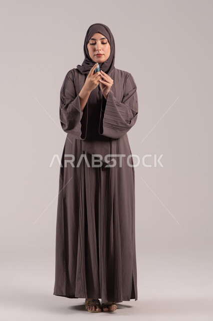 مشاكل في الجهاز التنفسي, شابة سعودية مريضة، المحافظة على ممر هوائي منتظم، بورتريه لامرأة عربية خليجية اماراتية محجبة ترتدي عباءة تحمل بيدها جهاز الرذاذ الخاص بمرض الربو تقوم باستنشاق أدوية الكحة، صورة الجسم بالطول الكامل، خلفية بنية