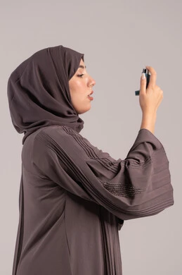 المحافظة على ممر هوائي منتظم, شابة سعودية مريضة، مشاكل في الجهاز التنفسي، بورتريه من الجانب لامرأة عربية خليجية اماراتية محجبة ترتدي عباءة تحمل بيدها جهاز الرذاذ الخاص بمرض الربو تقوم باستنشاق أدوية الكحة، خلفية بنية