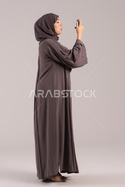 شابة سعودية مريضة، المحافظة على ممر هوائي منتظم، مشاكل في الجهاز التنفسي، بورتريه من الجانب لامرأة عربية خليجية اماراتية محجبة ترتدي عباءة تحمل بيدها جهاز الرذاذ الخاص بمرض الربو تقوم باستنشاق أدوية الكحة، صورة الجسم بالطول الكامل، خلفية بنية