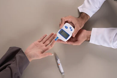 متابعة سكر الدم بالمنزل، بورتريه مقرب ليد ممرض يرتدي اليونيفورم يقوم بفحص نسبة السكر في الدم لمريضة عربية خليجية إماراتية ترتدي العباءة، خدمات تمريض منزلي، فحص طبي سريع ومباشر، خلفية بنية