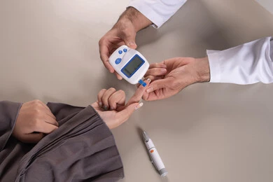 جهاز مراقبة سكر الدم الشخصي للمنزل، بورتريه مقرب ليد ممرض يرتدي اليونيفورم يقوم بفحص نسبة السكر في الدم لمريضة عربية خليجية إماراتية ترتدي العباءة، تمريض منزلي متكامل، متابعة صحية وفحص سريع، خلفية بنية