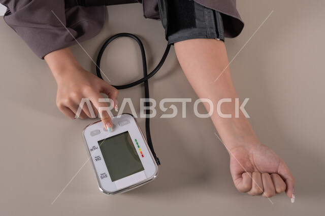 Regular follow-up for blood tests, care and attention to maintaining health, a close-up portrait of the hand of an Arab Gulf Emirati woman wearing an abaya measuring blood pressure using an electronic scale, the concept of medicine and healthcare, brown background.