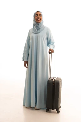 التجهيز للسفرات الممتعة, الاستمتاع بالرحلات السياحية في وقت العطلة, بورتريه لامرأة عربية خليجية سعودية محجبة ترتدي العباءة تمسك بيدها حقيبة سفر رمادية، النظر بإيماءات السعادة والسرور، الجسم بالطول الكامل, خلفية بيضاء