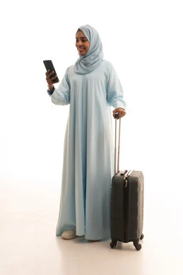 The use of devices, technologies, and tourism applications, the feeling of joy in the advanced services of tourism and travel programs, a portrait of a Saudi Arabian Gulf woman wearing a hijab and an abaya, holding a travel bag and a mobile phone in her hand, looking at the camera with gestures of happiness, full-body image, white background.