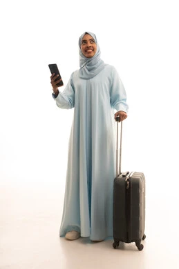 The feeling of joy in the advanced services of tourism and travel programs, the use of devices, technologies, and tourism applications, a portrait of a Saudi Arabian Gulf woman wearing a hijab and an abaya holding a travel bag and a mobile phone, looking at the camera with gestures of happiness, full-body image, white background.
