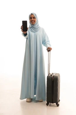 قضاء رحلة ممتعة خارج البلاد, استخدام جهاز تقني حديث متطور, بورتريه لامرأة عربية خليجية سعودية محجبة ترتدي العباءة تمسك بيدها حقيبة السفر وتنظر الى الكاميرا, عرض شاشة سوداء فارغة عبر الهاتف المحمول, صورة للجسم بالطول الكامل, خلفية بيضاء