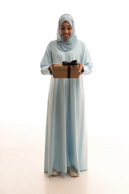 فرحة الحصول على الهدية والتهنئة، أعياد ومناسبات ومفاجآت سعيدة, النظر  بإيماءات السرورو السعادة ، بورتريه لامرأة عربية سعودية خليجية محجبة ترتدي عباءة تحمل بيديها علبة مغلفة، صورة الجسم بالطول الكامل، خلفية بيضاء