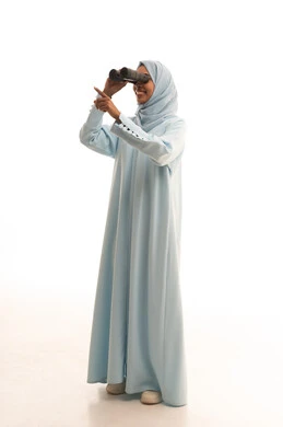 الاشارة الى شيء ما، رؤية الاشياء البعيدة عبر التلسكوب، التخطيط المستقبلي للأهداف و الطموحات، حب الاستكشاف والمغامرة، بورتريه لامرأة عربية خليجية سعودية محجبة ترتدي العباءة و الحجاب تنظر من خلال منظار ثنائي العينين, الجسم بالطول الكامل، خلفيه بيضاء