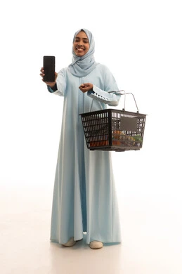 عرض شاشة سوداء فارغة، إيماءات السعادة والسرور، الاستمتاع بالتسوق في المولات والمراكز التجارية، شراء المستلزمات والحاجيات الضرورية، بورتريه لامرأة عربية خليجية سعودية محجبة تحمل بيدها الهاتف المحمول و سلة التسوق، تقديم خدمات وتطبيقات الموبايل، الجسم بالطول الكامل، خلفية بيضاء