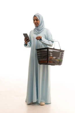 Chatting with family and friends, buying necessities and essential items, using modern technological devices, a portrait of a Saudi Arabian Gulf woman wearing a hijab holding a mobile phone and a shopping basket while browsing social media, full-body image, white background.