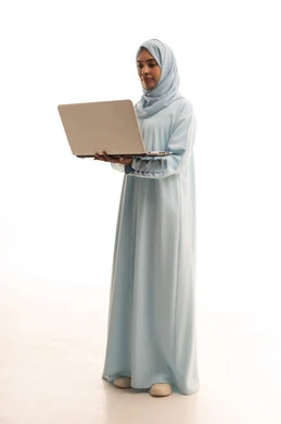 إنجاز المهام المطلوبة عبر اللابتوب، استخدام الأجهزة الحديثة والمتطورة، ادارة الأعمال عن بعد، بورتريه لامرأة عربية خليجية سعودية ترتدي العباءة والحجاب تقوم باستخدام الحاسب المحمول، دمج التكنولوجيا بالعمل، الجسم بالطول الكامل، خلفية بيضاء