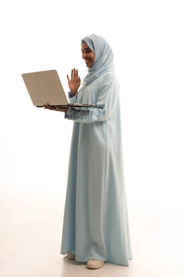 اجراء مكالمة فيديو عبر الانترنت، رفع كف اليد بتعابير السلام و التحية، ادارة الأعمال عن بعد، بورتريه لامرأة  عربية خليجية سعودية ترتدي العباءة والحجاب تقوم باستخدام الحاسب المحمول، دمج التكنولوجيا بالعمل، إنجاز المهام المطلوبة عبر اللابتوب، الجسم بالطول الكامل، خلفية بيضاء