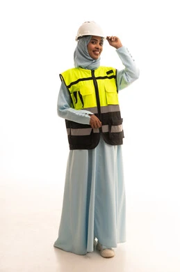 العمل في القطاع الهندسي، مخططات و مشاريع اعمارية، مهن ووظائف نسائية هندسية، بورتريه لامرأة عربية خليجية سعودية محجبة ترتدي خوذة وسترة الحماية تنظر الى الكاميرا بايماءات السعادة و السرور، الاشراف على العمل، صورة لكامل الجسم، خلفية بيضاء