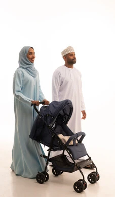 Care and attention to children, an Omani family spending quality time, the importance of parents and their essential role in raising children, a portrait of an Arab Gulf Omani woman wearing a hijab and abaya standing with her husband pulling a baby stroller, going on a family outing, white background.