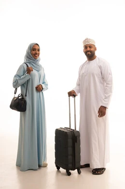 انتظار موعد الطائرة، الاستعداد لقضاء رحلة صيفية ممتعة، مفهوم السياحة والقيام بالجولات الترفيهية في وقت الإجازة، بورتريه لرجل عربي خليجي عماني يرتديالدشداشة و الكمة  يمسك بحقيبة الامتعة يقف برفقة زوجته، صورة الجسم بالطول الكامل، خلفية بيضاء
