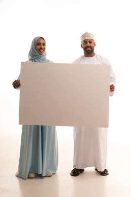 Gestures of happiness and joy, use of illustrative boards and educational tools, advertisements and marketing presentations, a portrait of an Arab Gulf Omani man wearing a dishdasha and kumma holding a large white board (empty mock-up) accompanied by his wife, full body length, white background.