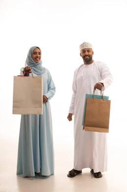 مشاركة الزوجين في إختيار المشتريات، الاستفادة من التخفيضات والعروض الموسمية، قضاء وقت ممتع بشراء الأغراض في الجمعة البيضاء، بورتريه لزوجان عربيان خليجيان عمانيان يحملان أكياس التسوق الملونة، تسوق المنتجات العالمية ، الطول الكامل للجسم، خلفية بيضاء