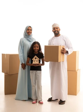 Gestures of happiness and joy at owning a new home, the concept of owning a new home and family stability, a portrait of an Arab Gulf Omani man wearing a dishdasha and a kumma holding a cardboard box in his hands accompanied by his wife and daughter, a girl holding a wooden model of a small house, white background.