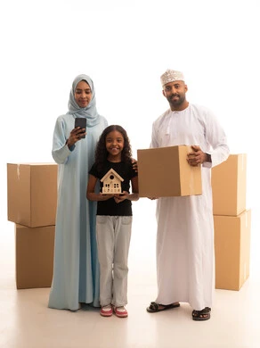 تصفح مواقع التواصل الاجتماعي عبر الهاتف المحمول ، مفهوم امتلاك سكن جديد والاستقرار العائلي، بورتريه لرجل عربي خليجي عماني يرتدي الدشداشة و الكمة يحمل بيدية صندوق كرتوني برفقة زوجته و ابنته، بنت تحمل مجسم خشبي لبيت صغير ، ايماءات السعادة والسرور بامتلاك سكن جديد