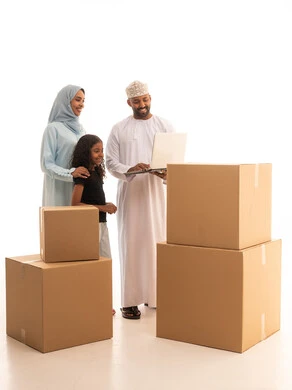 Moving to a new home, boxes and cardboard containers, using modern technologies, owning a new residence and family stability, a portrait of an Arab Gulf Omani man wearing a dishdasha and kumma using a laptop with his wife and daughter, full-body, white background.